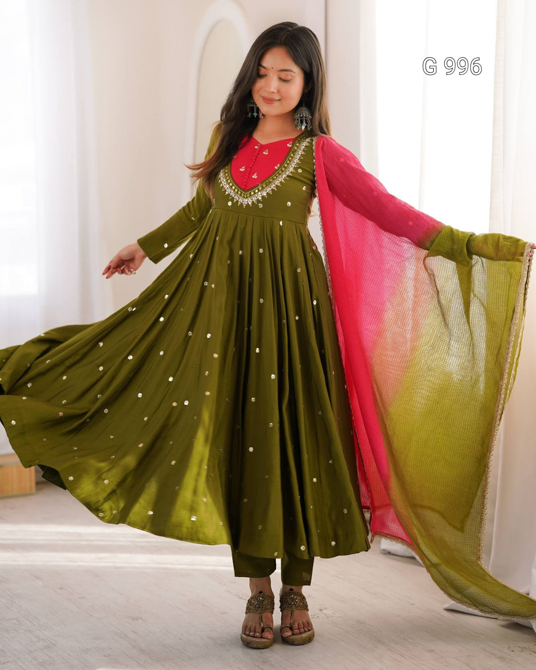 A woman modeling a traditional Indian outfit featuring an olive green Anarkali-style dress with gold dot embellishments, a bright pink inner neckline, and a matching pink and olive green gradient dupatta, set against a bright indoor background.