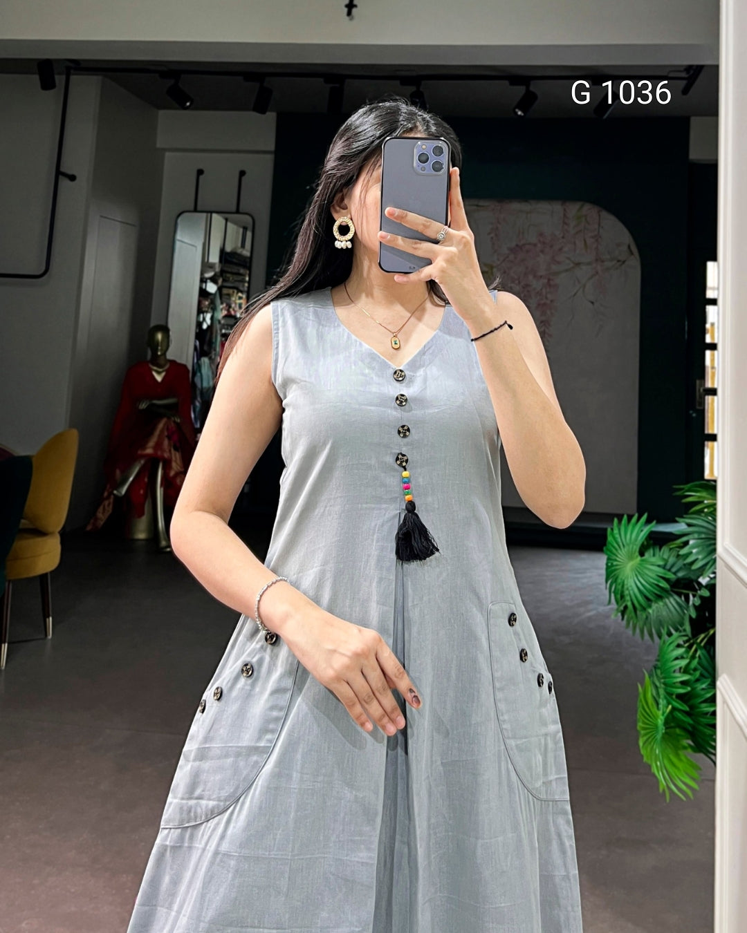 A woman takes a mirror selfie wearing a sleeveless light gray linen dress with a V-neckline, decorative buttons, and a tassel detail. The background shows a boutique interior with a mannequin and a plant.