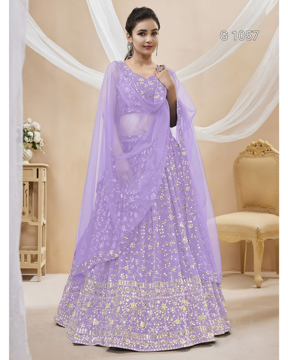 A woman models a lavender purple lehenga choli set with gold mirror work and embroidery, featuring a sheer dupatta, in a decorated indoor studio setting.