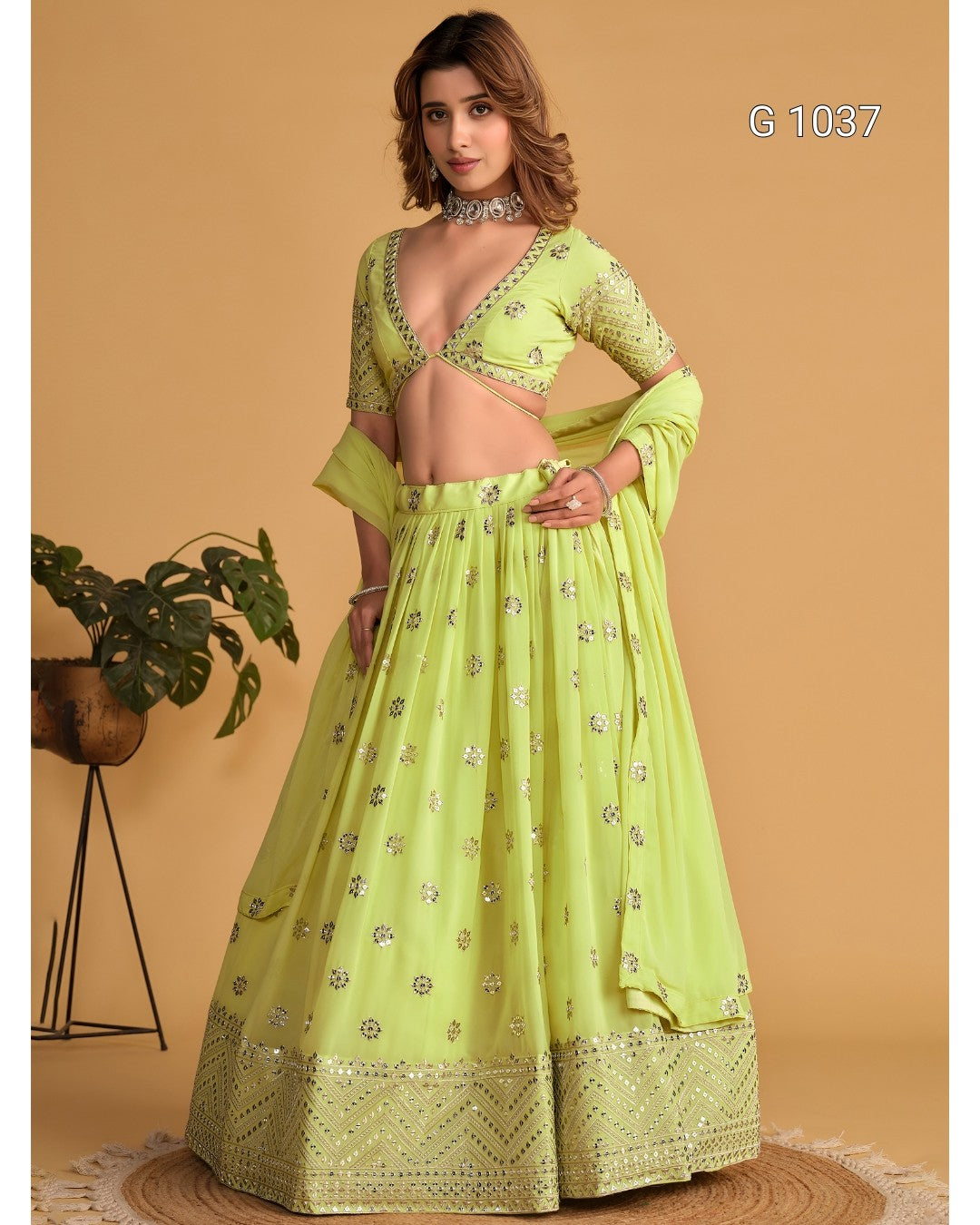 A woman models a light lime green ethnic lehenga choli set with silver mirror work embroidery, set against a warm beige background with a potted plant on the side.
