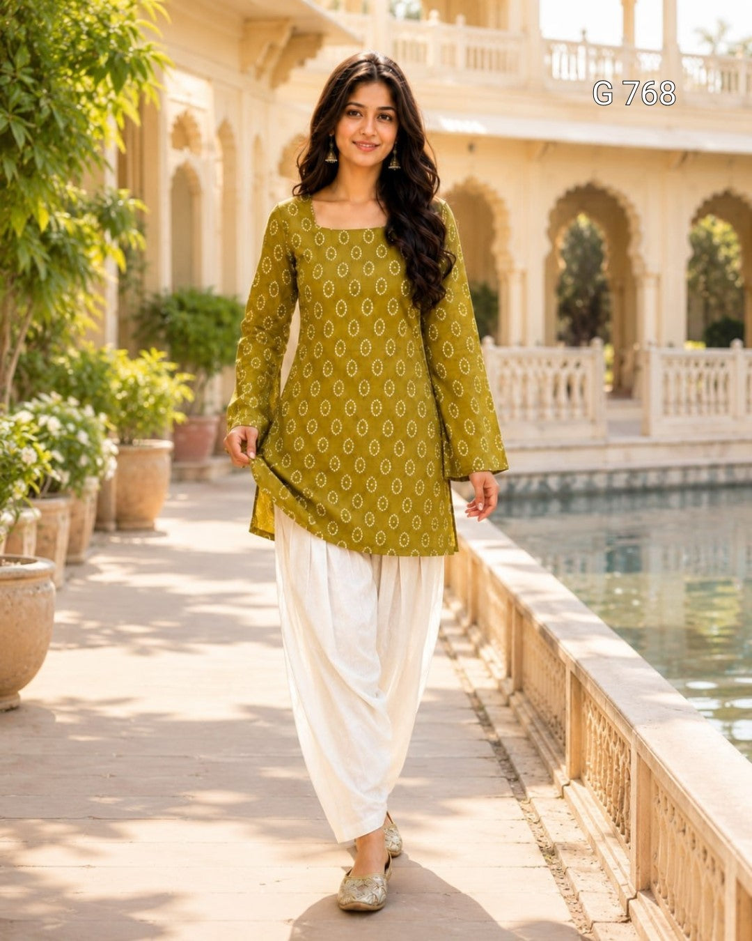Woman in a green patterned top and white pants standing in a scenic outdoor setting.