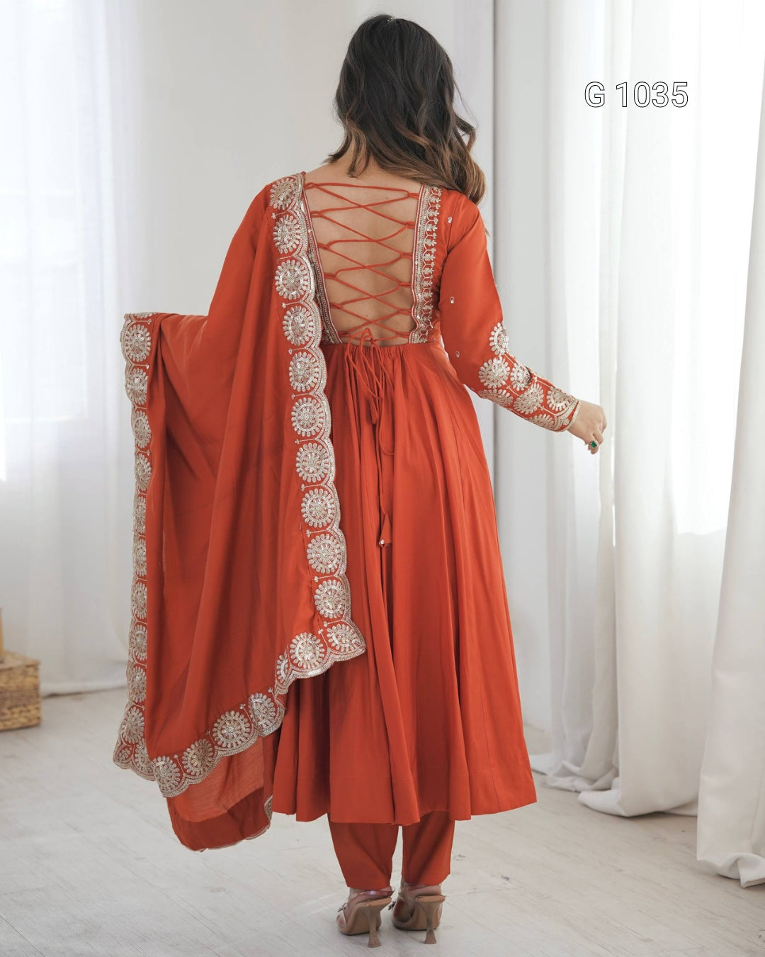 A woman shown from the back wearing a rust-orange traditional outfit with a matching dupatta, featuring intricate silver embroidery and a criss-cross lace-up back. The setting is a bright room with white curtains.