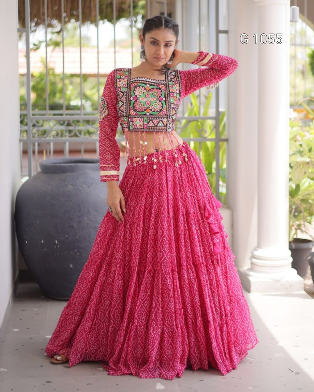 A woman models a bright pink traditional Indian outfit with a heavily embroidered floral crop top and a matching flared skirt, standing on a porch with columns and potted plants in the background.