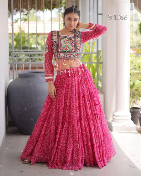 A woman models a bright pink traditional Indian outfit with a heavily embroidered floral crop top and a matching flared skirt, standing on a porch with columns and potted plants in the background.