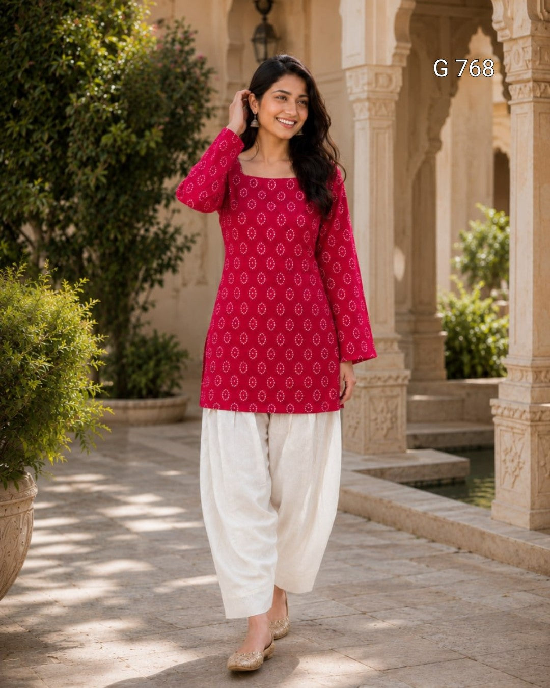Woman in a red patterned top and white pants standing in an outdoor setting with architectural elements.