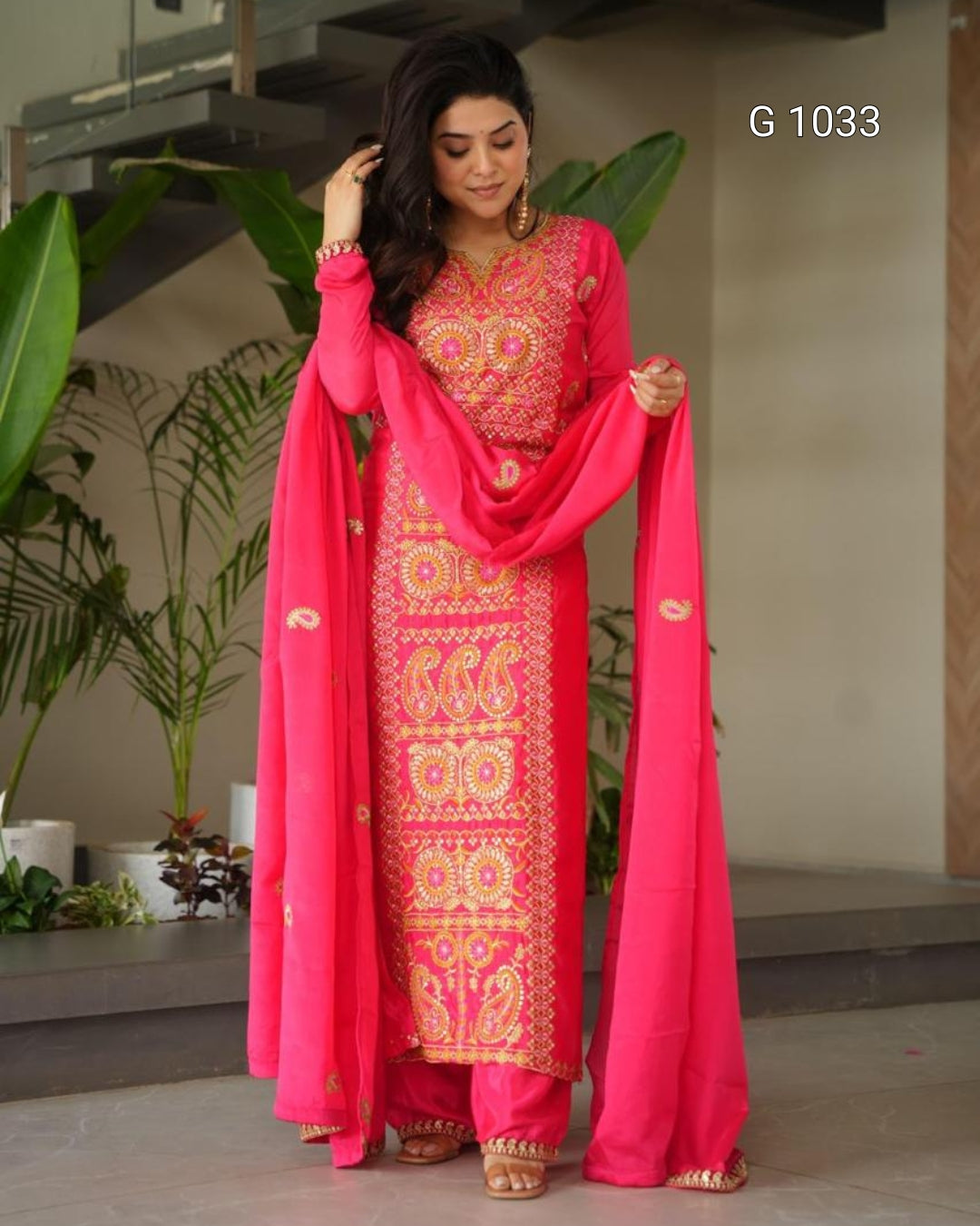 A woman modeling a bright pink Indian ethnic salwar kameez suit with intricate gold embroidery and a matching dupatta, standing in an indoor setting with plants in the background.