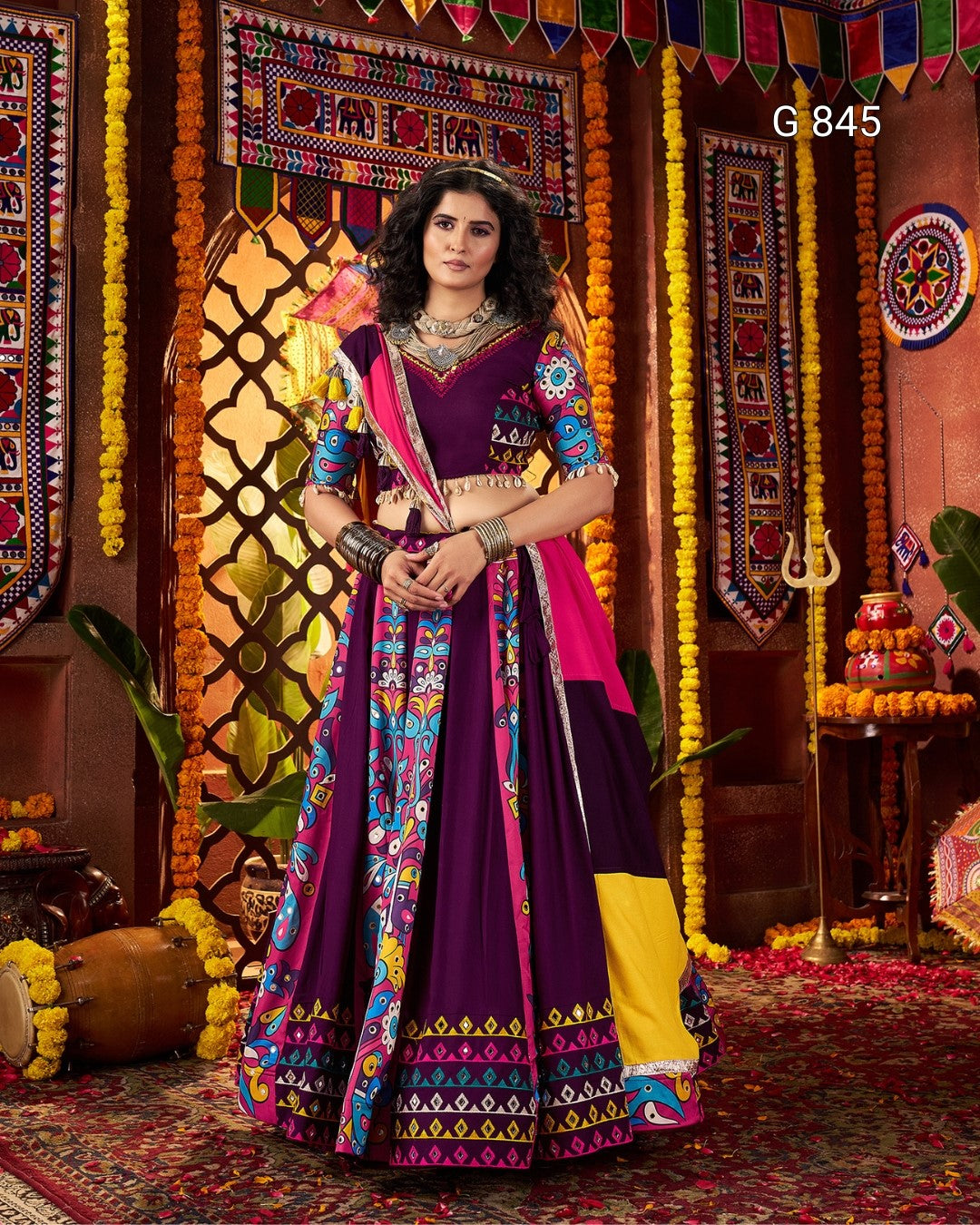 Woman in a colorful traditional outfit standing in front of decorative walls with patterns and flowers.