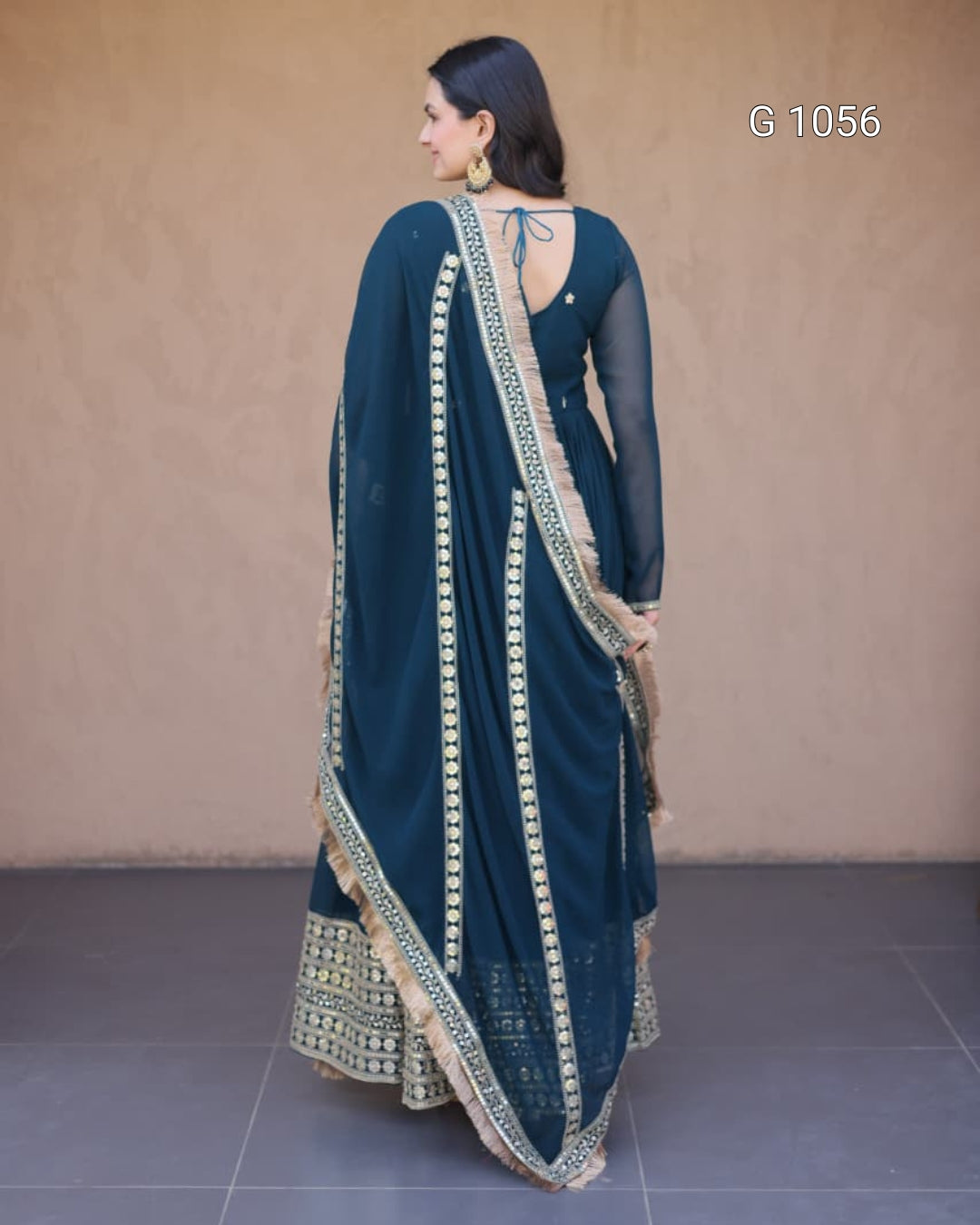 A woman shown from the back wearing a dark teal blue saree with beige and gold border patterns, standing against a plain beige wall.