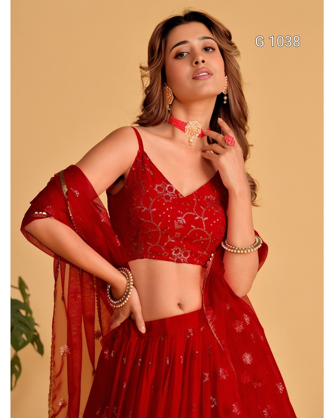 A woman modeling a red embroidered traditional outfit with a cropped embroidered blouse, matching skirt, and draped dupatta against a warm beige studio background.