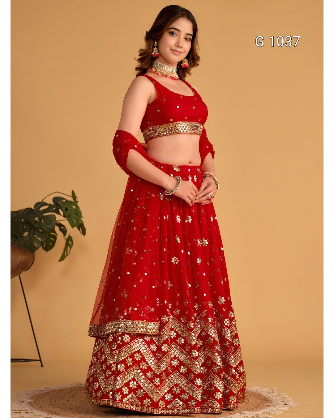 A woman modeling a red and gold embroidered traditional Indian lehenga choli dress against a warm beige studio background. The outfit features a red sleeveless blouse with gold embellishments, a matching red flared skirt with ornate gold border detailing, and red dupatta sleeves with gold floral embroidery.