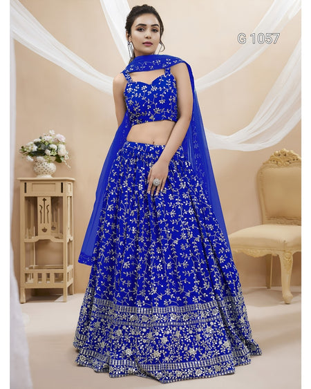 A woman models a royal blue lehenga choli set with intricate silver floral embroidery and a matching sheer dupatta, set against a warm beige background with draped white fabric and a decorative chair.