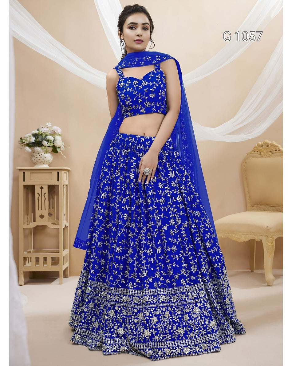 A woman models a royal blue lehenga choli set with intricate silver floral embroidery and a matching sheer dupatta, set against a warm beige background with draped white fabric and a decorative chair.