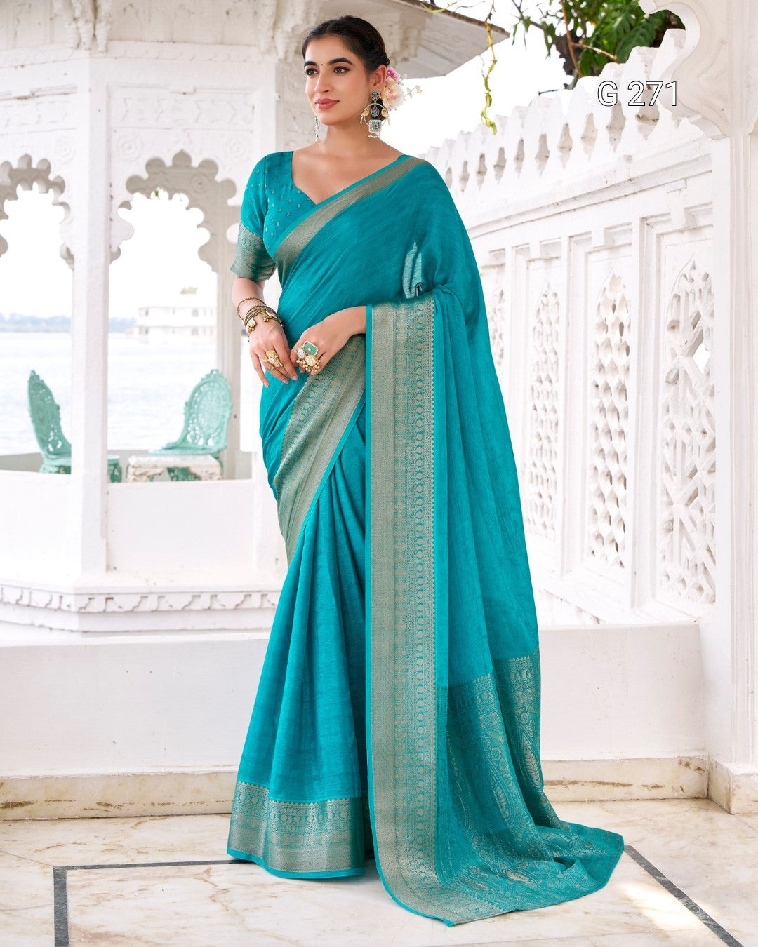 A woman models a teal green saree with a matching blouse in a white architectural pavilion. The saree features a wide border with gold and silver zari embroidery.