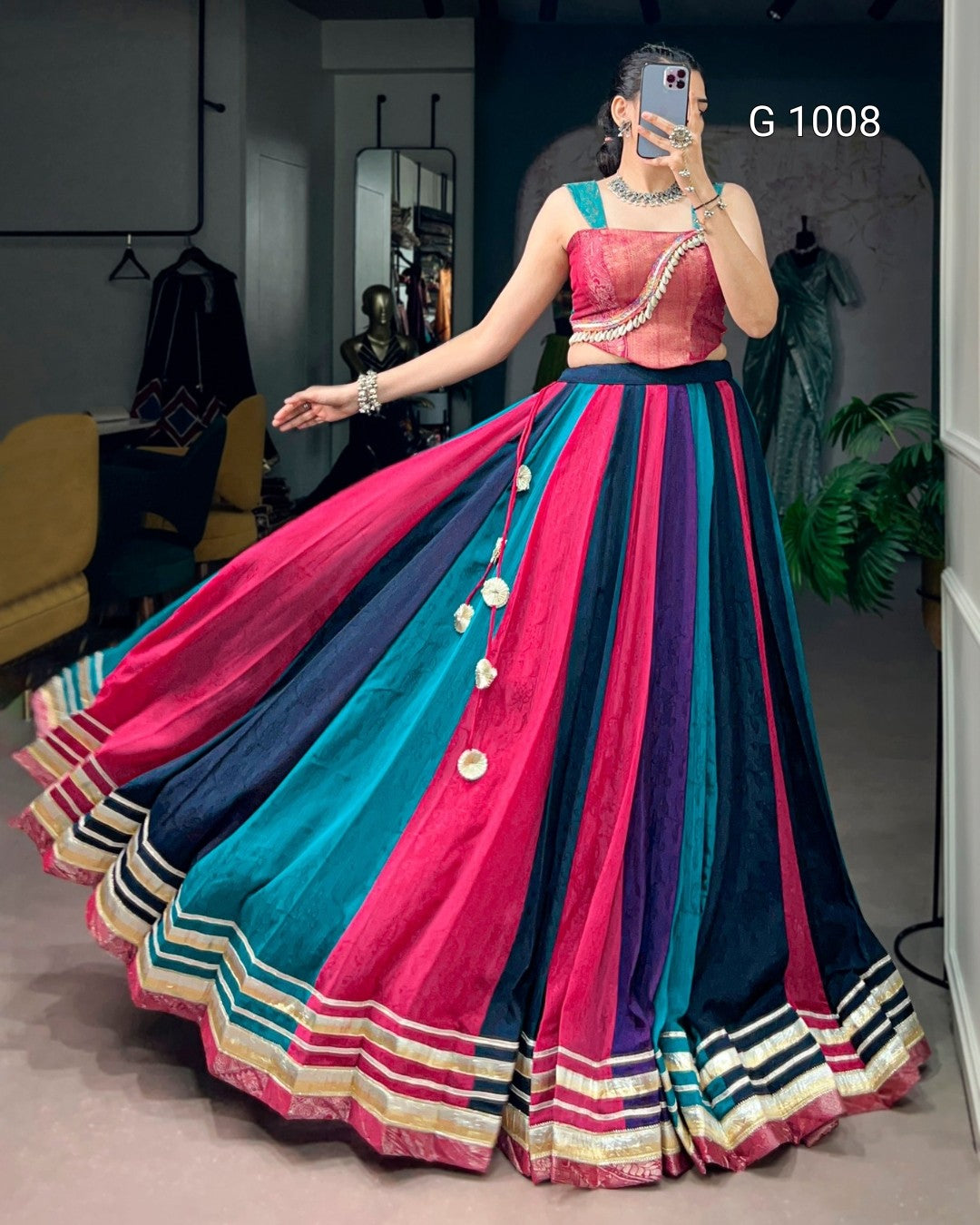 A woman takes a mirror selfie in a boutique wearing a traditional Indian lehenga choli. The dress features a bright pink and teal embroidered bodice with a black waistband, and a voluminous skirt with vertical stripes of pink, teal, black, and gold with decorative coin-like embellishments.