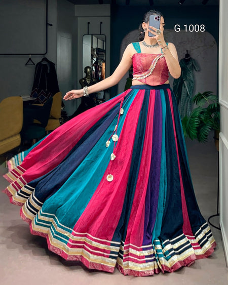 A woman takes a mirror selfie in a boutique wearing a traditional Indian lehenga choli. The dress features a bright pink and teal embroidered bodice with a black waistband, and a voluminous skirt with vertical stripes of pink, teal, black, and gold with decorative coin-like embellishments.