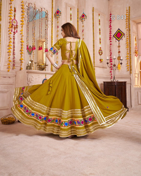 Woman in a green traditional outfit with colorful embroidery in a decorated room.