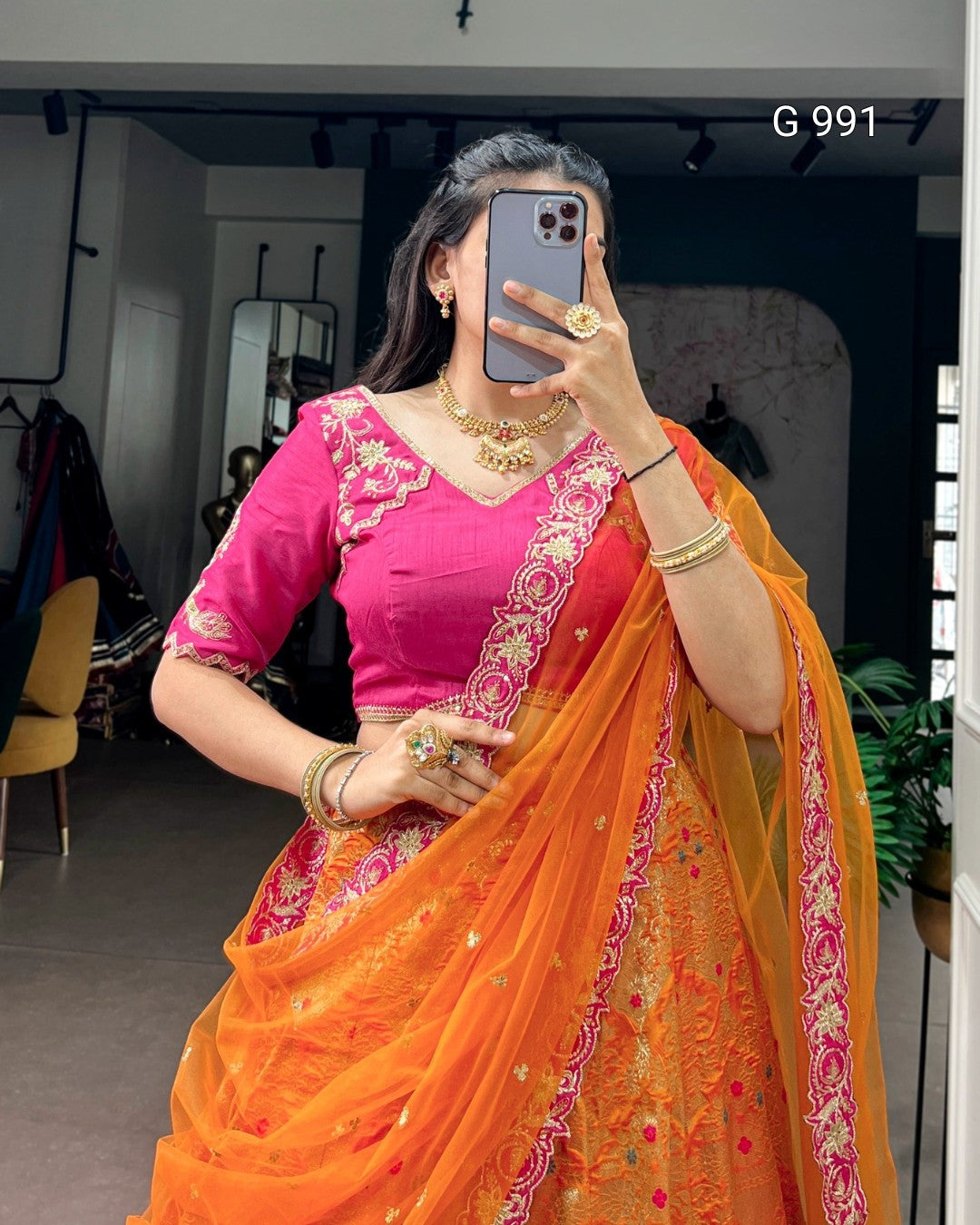 A woman takes a mirror selfie wearing a traditional Indian outfit consisting of a bright pink embroidered blouse and an orange saree with pink and gold floral border details. She is wearing gold jewelry and holding a dark gray smartphone.