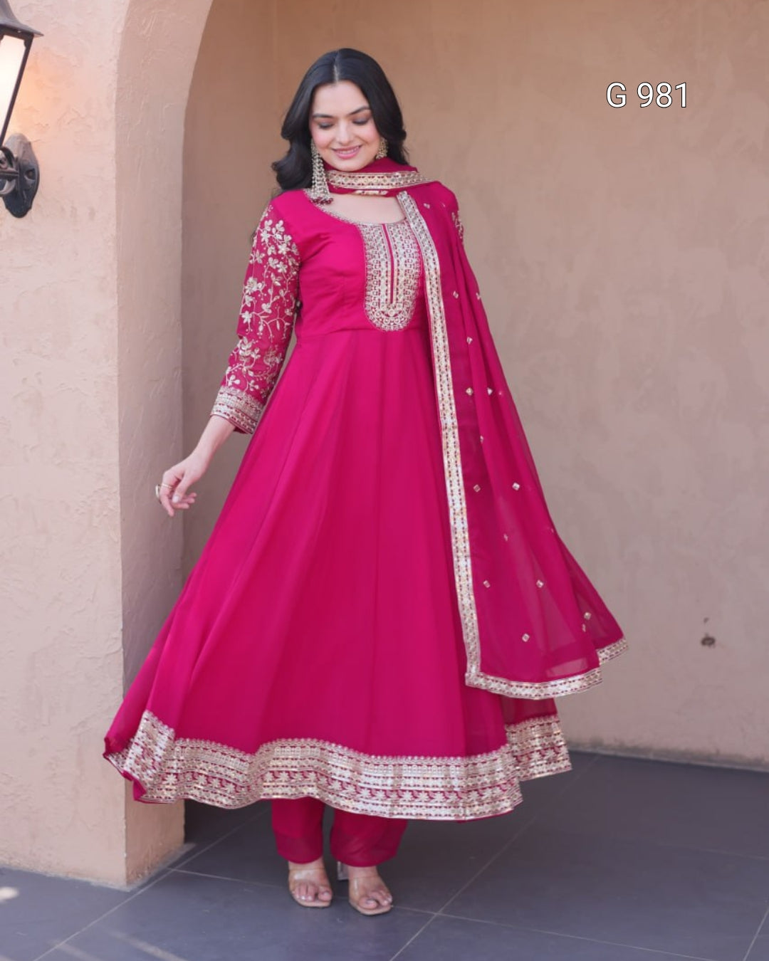 A woman modeling a bright pink traditional Anarkali suit with gold embroidery, matching pants, and a dupatta, standing against a beige stucco wall.