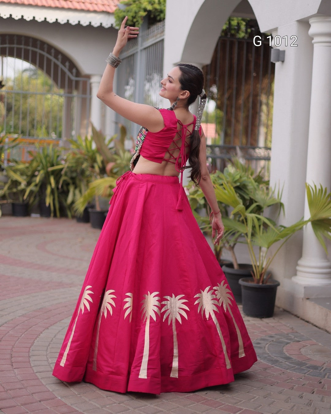 A woman modeling a bright pink traditional Indian outfit with a flared skirt featuring cream palm tree prints, set in front of a white arched building with potted plants.