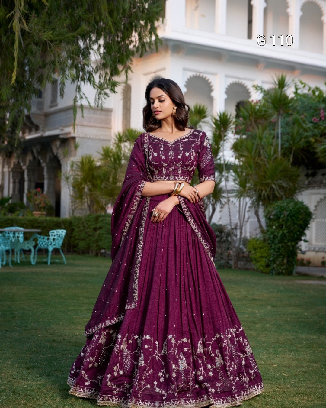 Woman in a purple traditional outfit standing outdoors with greenery and a building in the background.