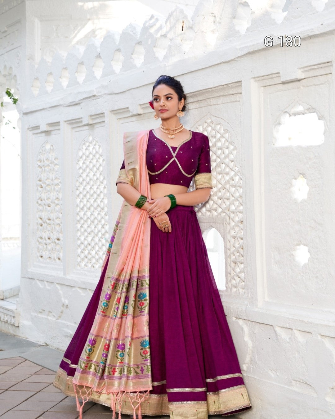 Woman in a purple traditional outfit with a pink dupatta against a white decorative wall.