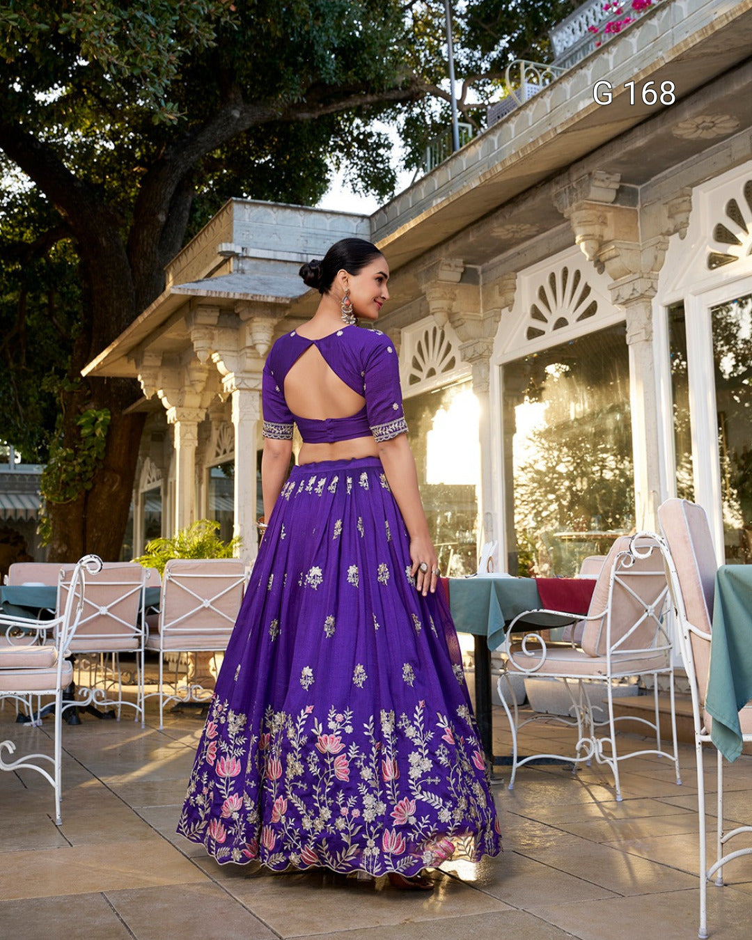 Woman in a purple embroidered traditional outfit standing outdoors with a building and trees in the background.