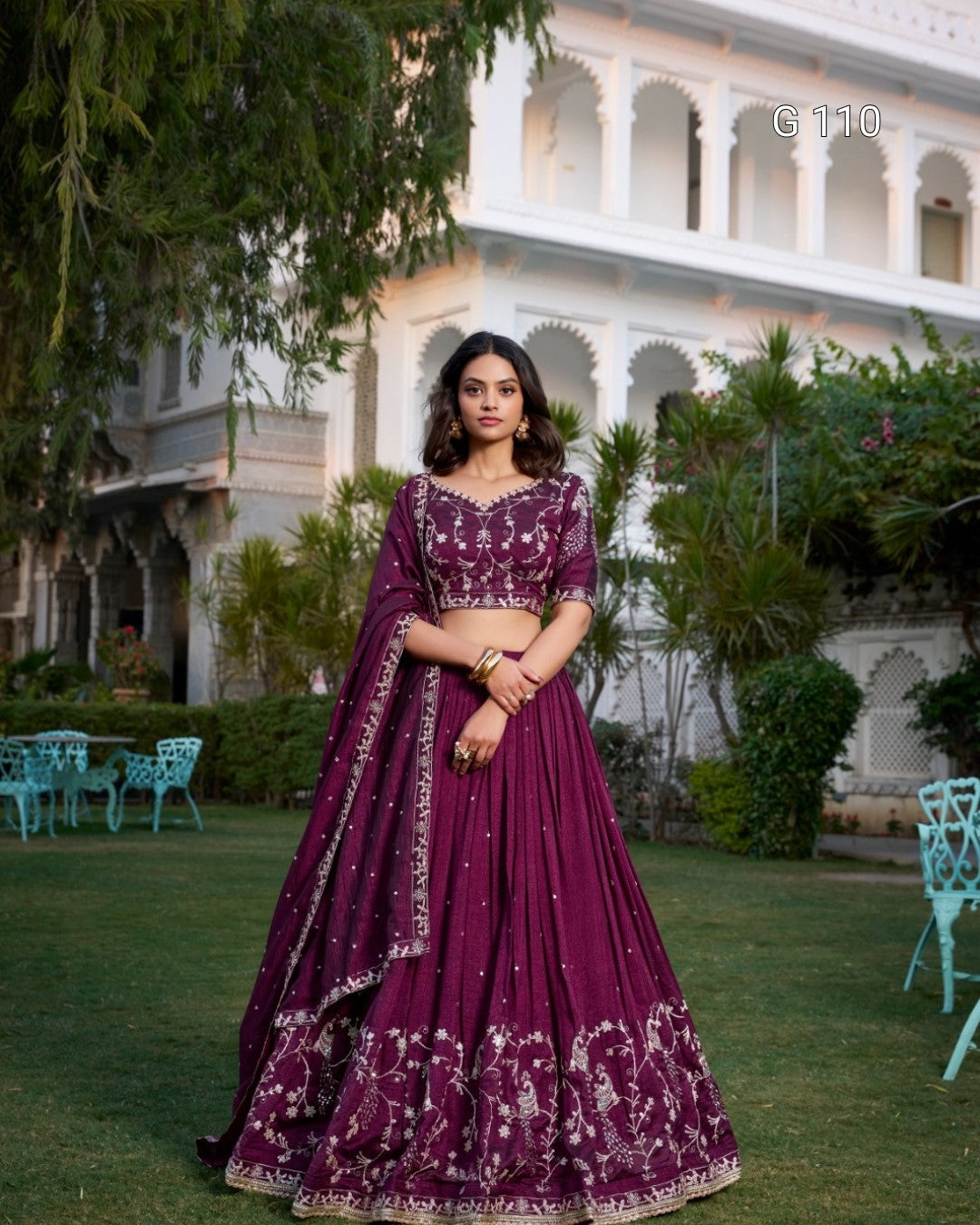 Woman in a purple traditional outfit standing in front of a building with greenery.