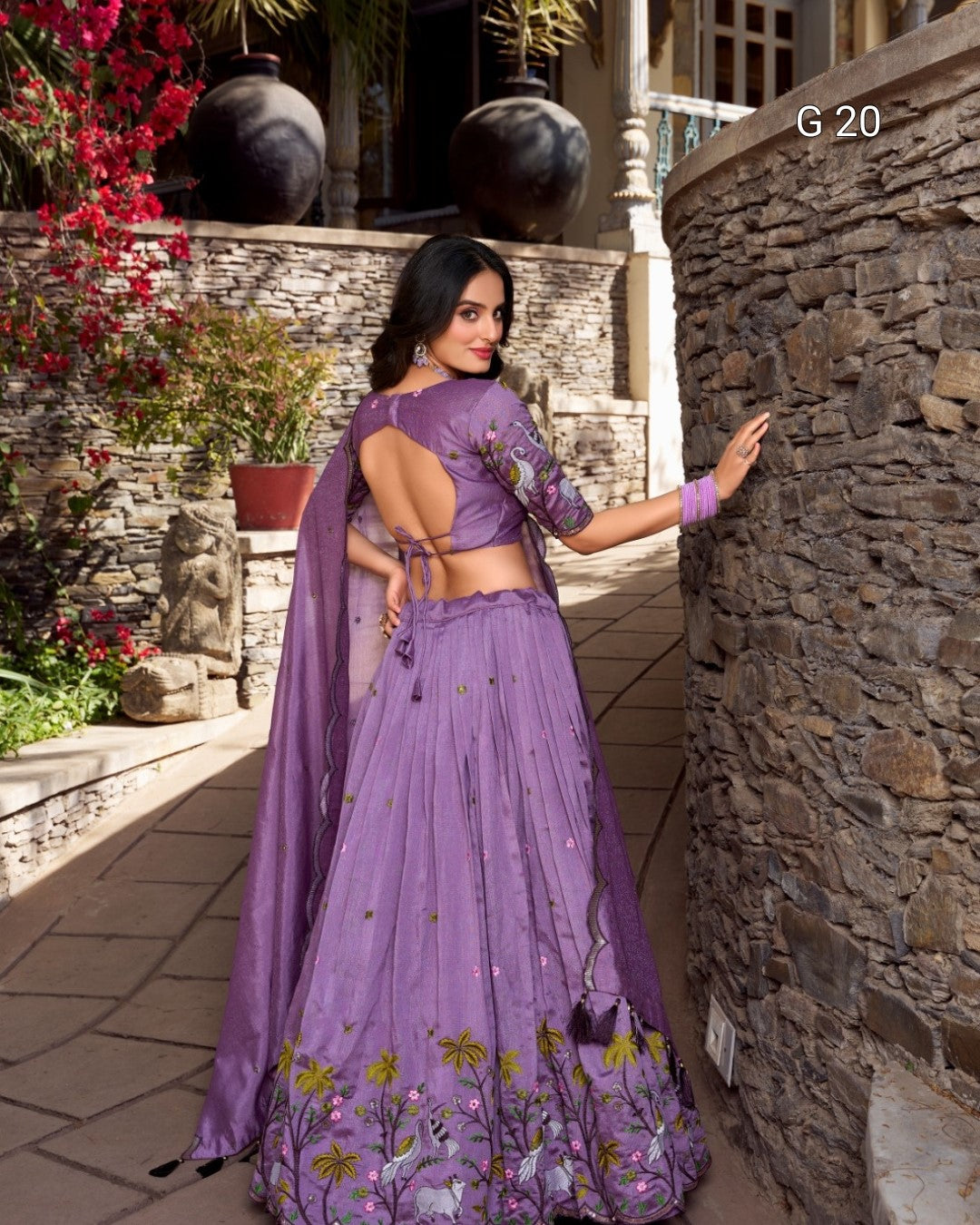 Woman in a purple traditional outfit standing in an outdoor setting with stone walls and plants.