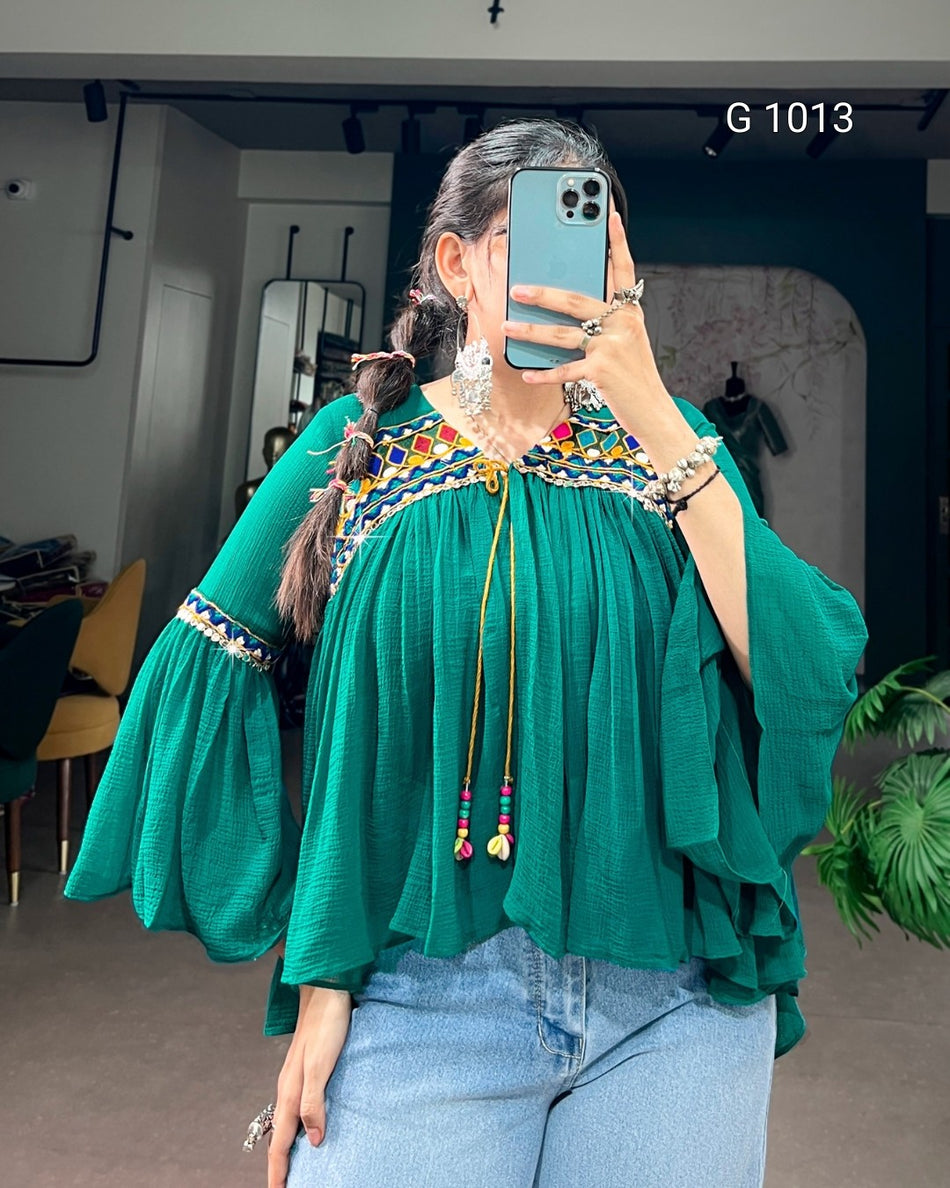 A woman takes a mirror selfie wearing a vibrant green bohemian-style blouse with tiered ruffle sleeves, a colorful embroidered yoke, and a beaded tassel tie at the neckline, paired with light blue jeans.