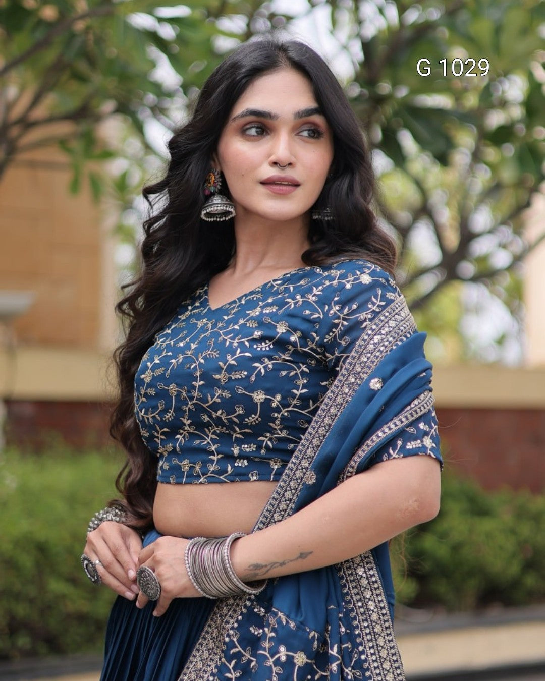 A woman wearing a blue embroidered saree with a matching crop top blouse, styled with silver jewelry, standing outdoors with a blurred garden background. The text 'G 1029' is visible in the top right corner.