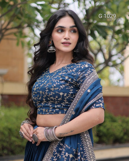 A woman wearing a blue embroidered saree with a matching crop top blouse, styled with silver jewelry, standing outdoors with a blurred garden background. The text 'G 1029' is visible in the top right corner.