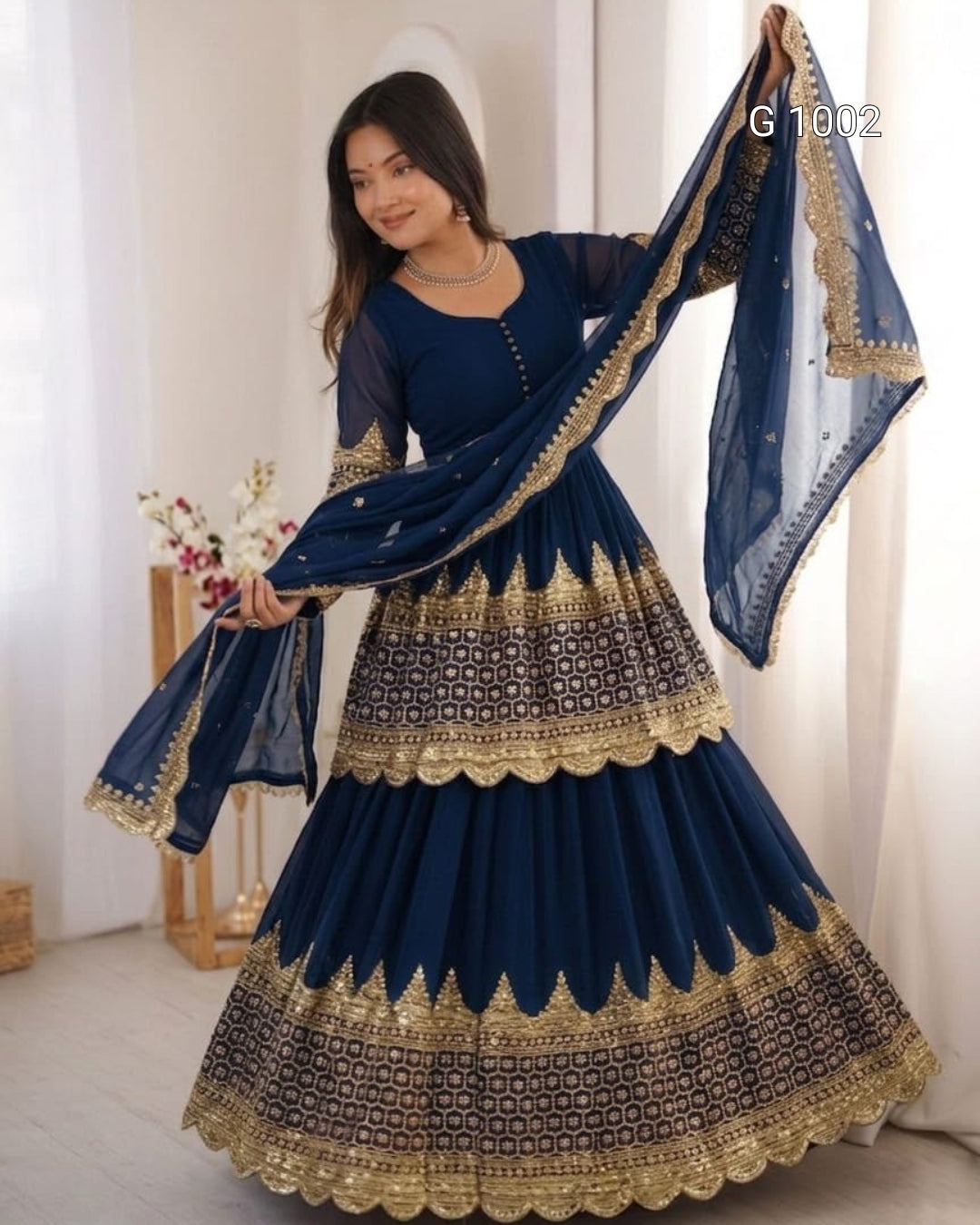 A woman modeling a navy blue traditional Anarkali gown with intricate gold embroidery and a matching dupatta, posed in a bright indoor setting.