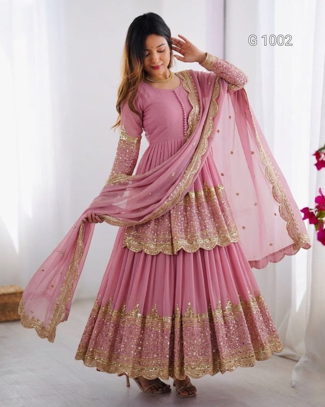 A woman modeling a pink traditional ethnic outfit with gold embroidery and a matching sheer dupatta, standing in a bright indoor setting with white curtains.