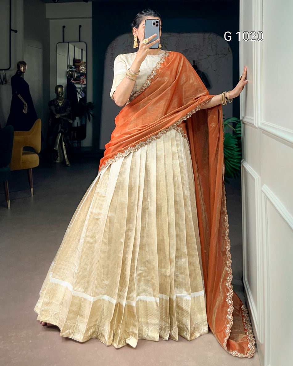 A woman takes a mirror selfie wearing a traditional Indian outfit consisting of a voluminous cream-colored pleated skirt (lehenga), a cream blouse with gold embroidery, and a draped orange dupatta with a gold embroidered border. The text 'G 1020' is visible in the top right corner.
