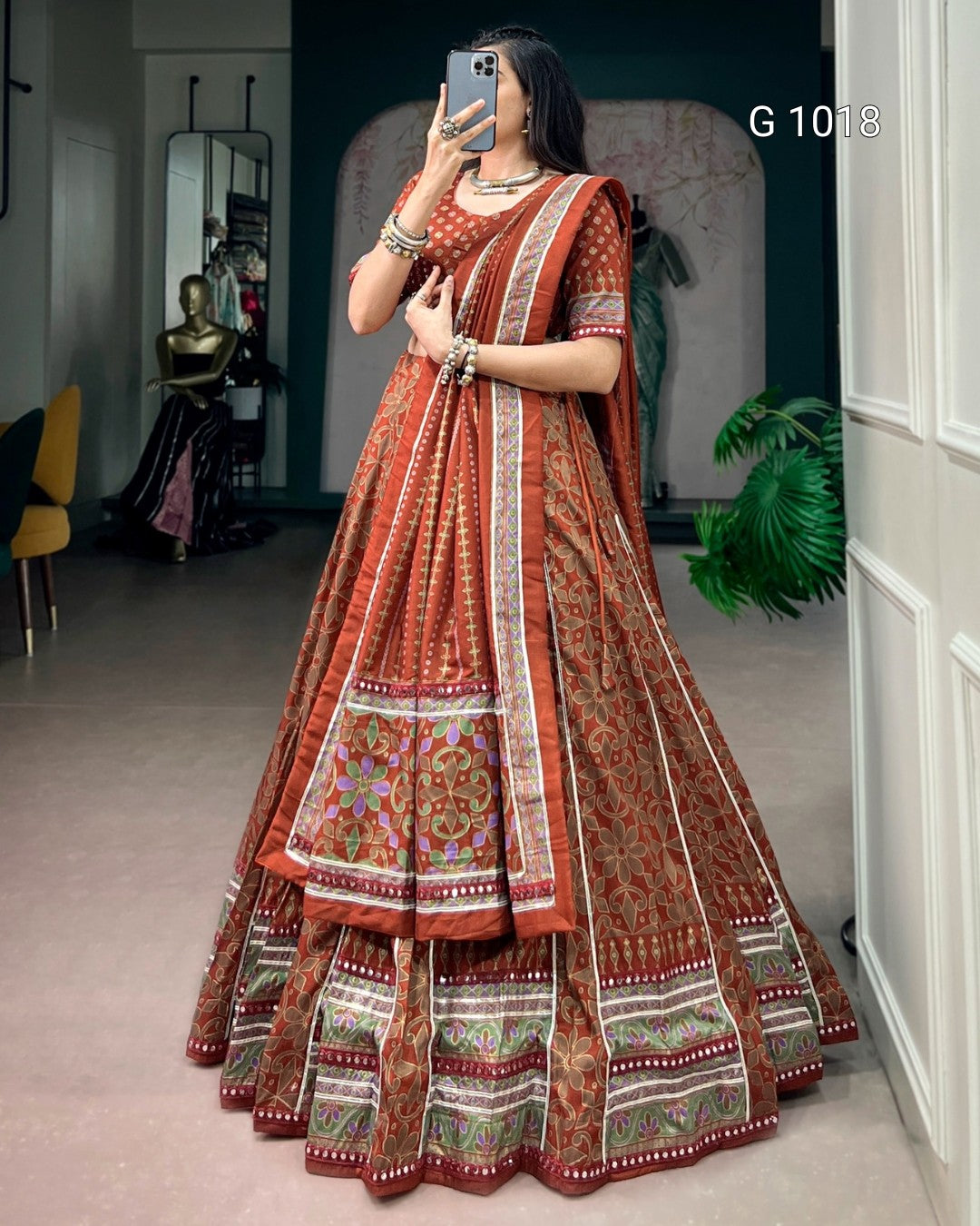 A woman takes a mirror selfie wearing a traditional Indian lehenga choli set in rust orange with intricate gold, green, and white border patterns. The setting is a boutique interior with a mannequin and plants visible in the background.