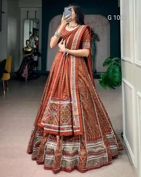 A woman takes a mirror selfie wearing a traditional Indian lehenga choli set in rust orange with intricate gold, green, and white border patterns. The setting is a boutique interior with a mannequin and plants visible in the background.