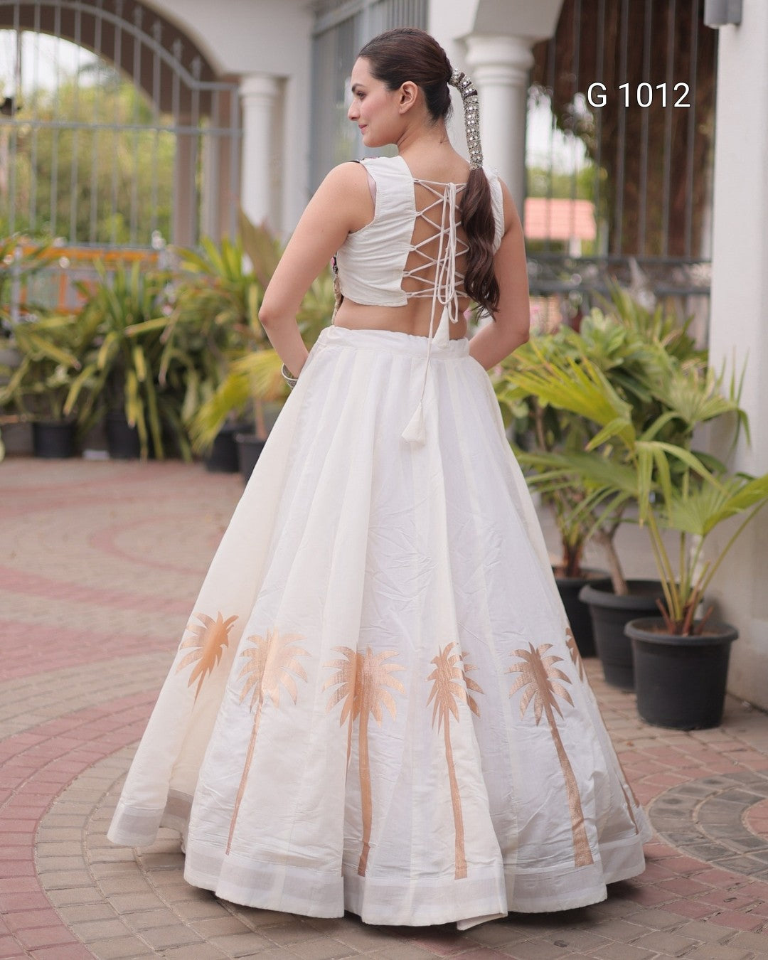 A woman wearing a white sleeveless lehenga choli dress with a lace-up open back and embroidered palm tree motifs near the bottom, photographed outdoors in front of a building with potted plants. The text 'G 1012' appears in the upper right.
