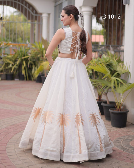 A woman wearing a white sleeveless lehenga choli dress with a lace-up open back and embroidered palm tree motifs near the bottom, photographed outdoors in front of a building with potted plants. The text 'G 1012' appears in the upper right.