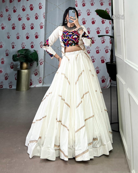 Woman in traditional outfit with colorful top and white skirt in a room with a patterned wall.