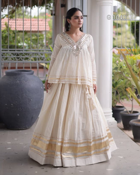 A woman modeling a traditional Indian outfit consisting of a cream-colored kurta and a matching flared skirt with gold and silver border details, set in a porch environment with potted plants.