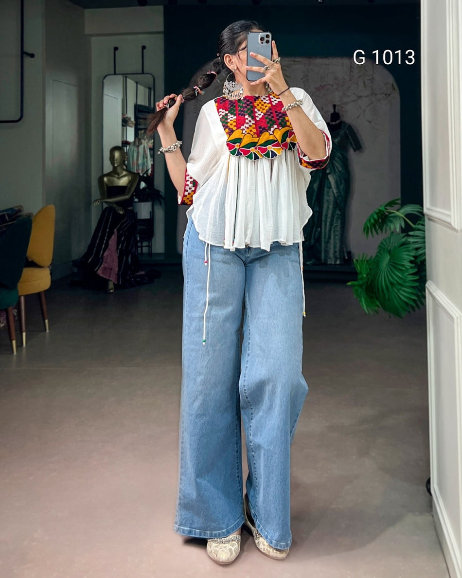 A woman takes a mirror selfie in a boutique showing a white blouse with a colorful geometric patterned yoke and wide-leg light blue jeans. The boutique interior has a neutral floor, a mannequin, and a green plant in the background.