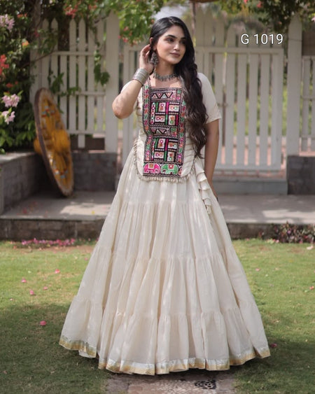 A woman wearing a cream-colored traditional Anarkali gown with a heavily embroidered multicolor bodice, standing outdoors on a grassy lawn in front of a white picket fence.