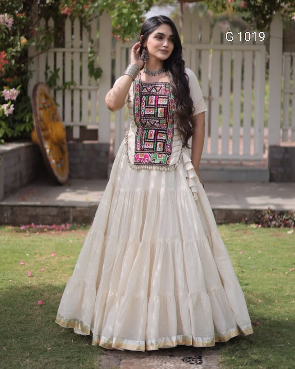 A woman wearing a cream-colored traditional Anarkali gown with a heavily embroidered multicolor bodice, standing outdoors on a grassy lawn in front of a white picket fence.