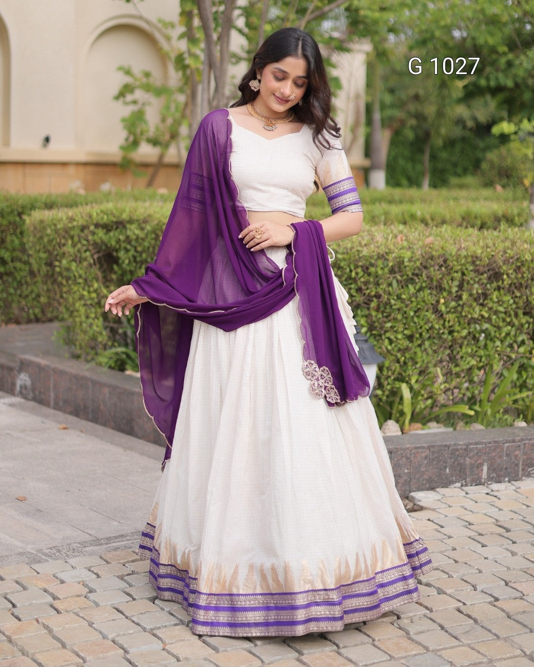 A woman modeling a traditional Indian outfit consisting of a white lehenga skirt with purple and gold borders, a white blouse with purple sleeve bands, and a purple dupatta, set against a garden background.