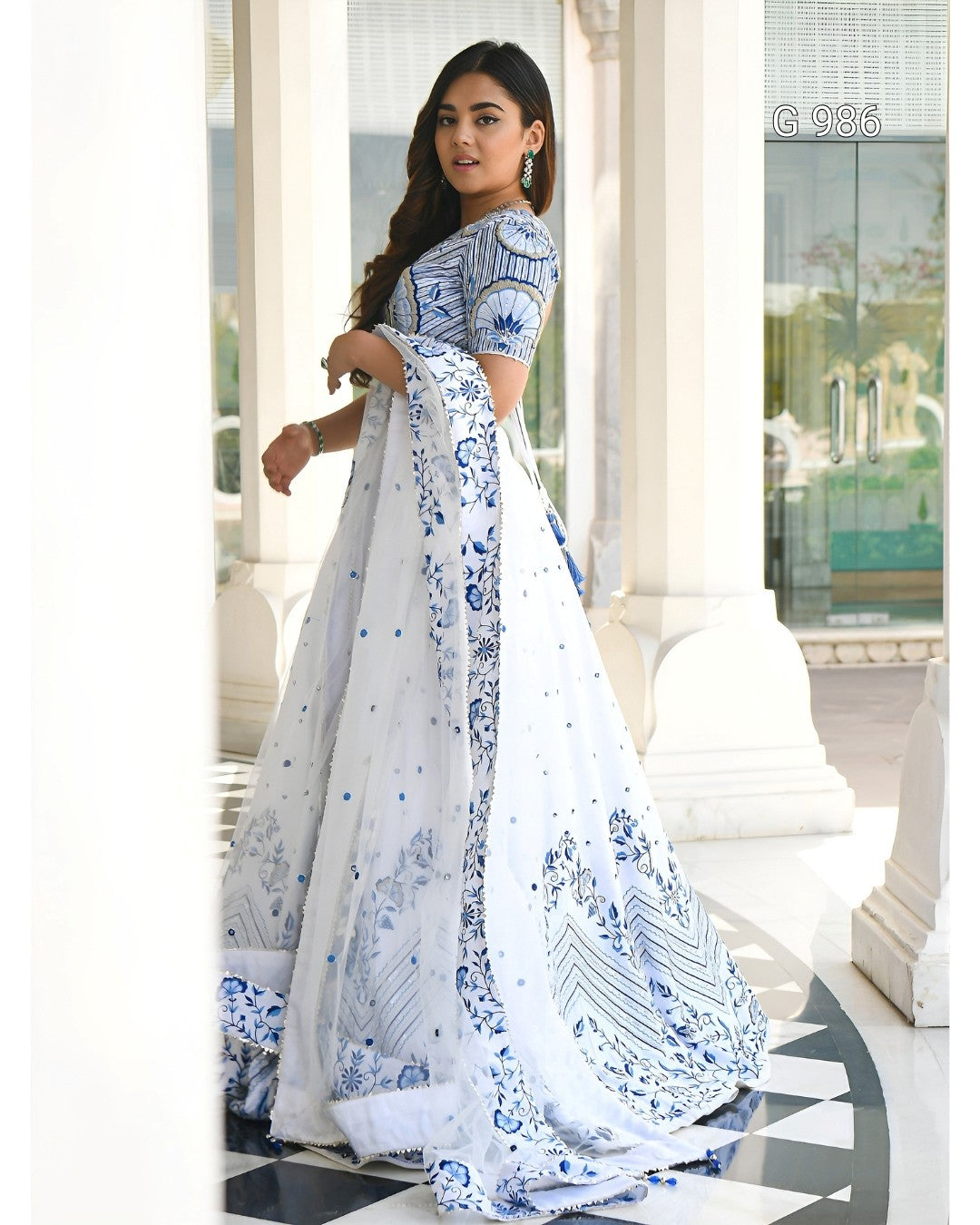 A woman modeling a white and blue printed traditional dress with a sheer dupatta in an elegant indoor setting with white pillars.