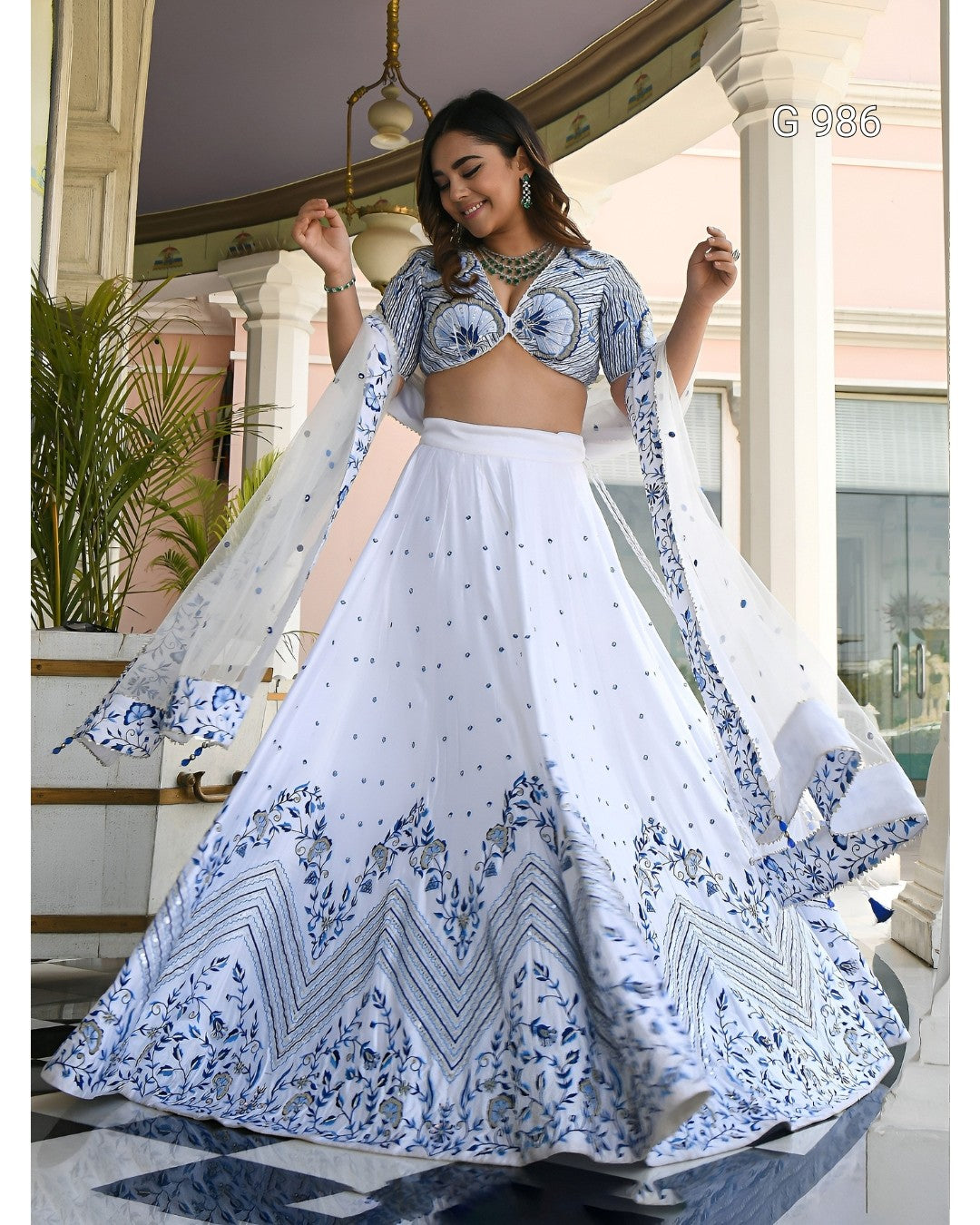 A woman modeling a white and blue printed traditional Indian lehenga choli set in an elegant indoor setting with pillars and plants. The outfit features a white flared skirt with intricate blue patterns, a matching printed blouse, and sheer dupatta panels.