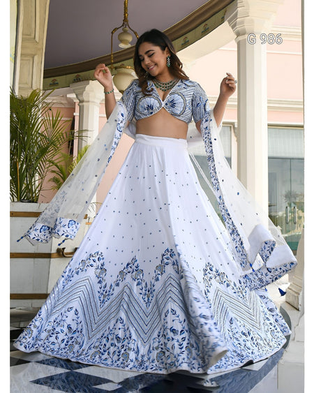 A woman modeling a white and blue printed traditional Indian lehenga choli set in an elegant indoor setting with pillars and plants. The outfit features a white flared skirt with intricate blue patterns, a matching printed blouse, and sheer dupatta panels.