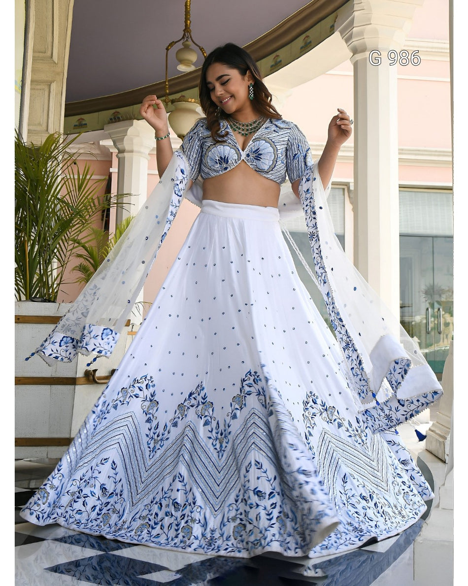 A woman modeling a white and blue printed traditional Indian lehenga choli set in an elegant indoor setting with pillars and plants. The outfit features a white flared skirt with intricate blue patterns, a matching printed blouse, and sheer dupatta panels.