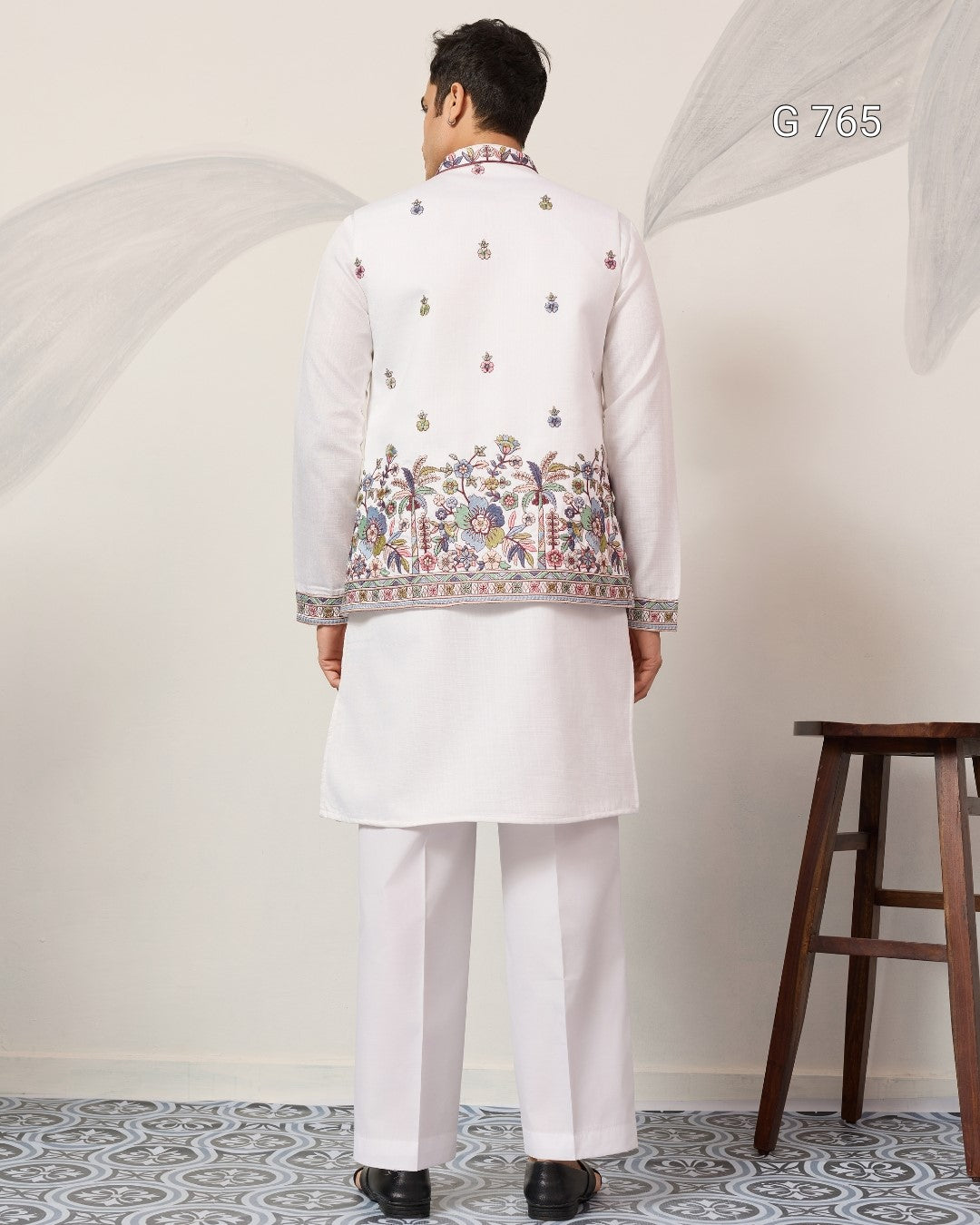 Person wearing a traditional embroidered jacket over a white kurta in a minimalistic setting.
