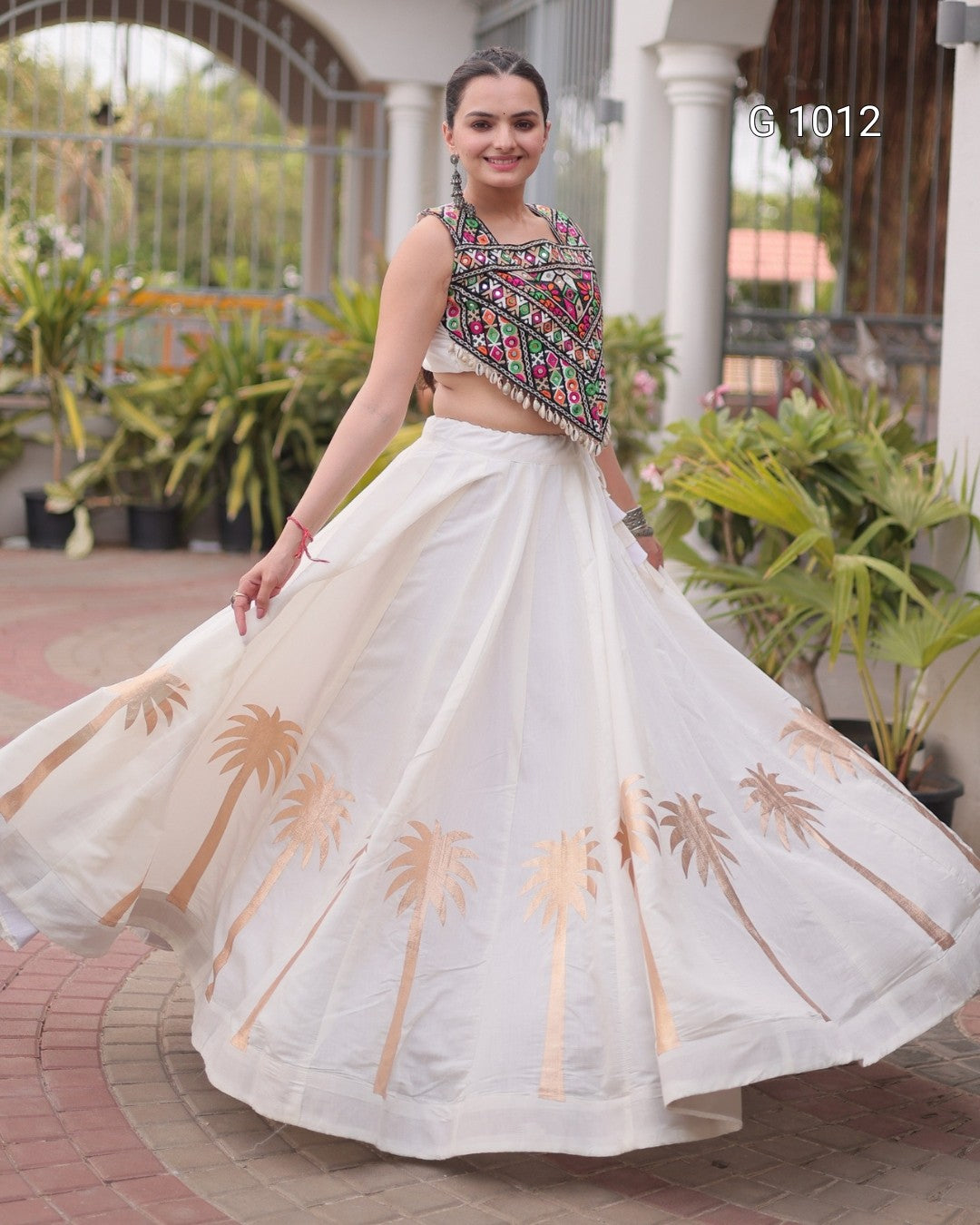 A woman wearing a white traditional lehenga choli outfit with embroidered details and gold palm tree prints, captured in a courtyard setting.